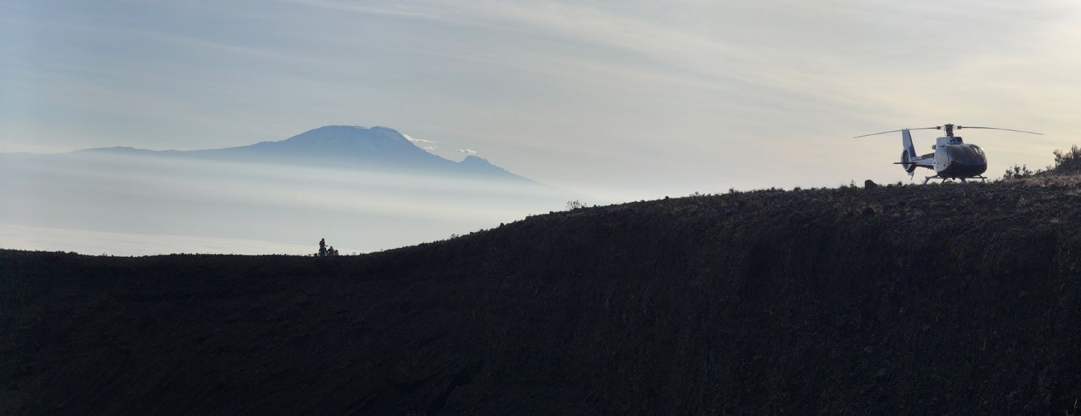 A helicopter parked on a ridge of Mount Meru with Kilimanjaro above the clouds